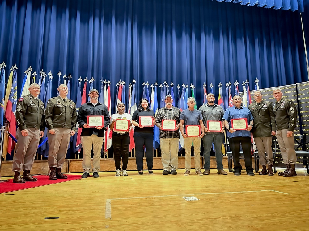 A formal awards ceremony taking place on a stage at Fort Campbell military installation, where several civilians are being recognized with certificates by military officers. A group of approximately eleven individuals stand in a line on a wooden stage in front of a blue curtain and a row of military and national flags. Two uniformed military officers flank a group of civilian recipients holding red-framed certificates.