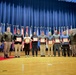 A formal awards ceremony taking place on a stage at Fort Campbell military installation, where several civilians are being recognized with certificates by military officers. A group of approximately eleven individuals stand in a line on a wooden stage in front of a blue curtain and a row of military and national flags. Two uniformed military officers flank a group of civilian recipients holding red-framed certificates.