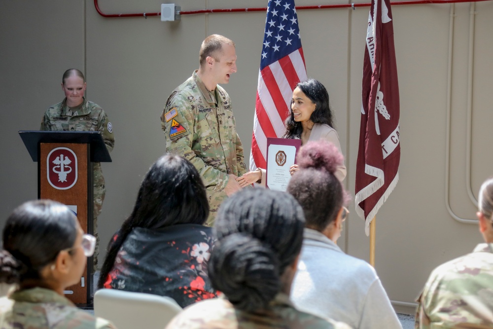A U.S. Army officer in camouflage uniform is presenting a certificate or award to a civilian woman during a formal military ceremony. A second uniformed soldier stands at a podium in the background. An audience of both military personnel and civilians is seated in the foreground.