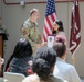 A U.S. Army officer in camouflage uniform is presenting a certificate or award to a civilian woman during a formal military ceremony. A second uniformed soldier stands at a podium in the background. An audience of both military personnel and civilians is seated in the foreground.