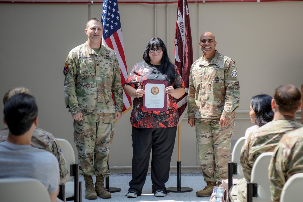 A formal military award ceremony is taking place indoors, with a civilian woman holding a framed certificate flanked by two U.S. Army soldiers in OCP (Operational Camouflage Pattern) uniforms. The setting includes an American flag and a maroon military unit guidon in the background. An audience of seated military personnel and civilians observes the presentation.
