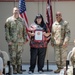 A formal military award ceremony is taking place indoors, with a civilian woman holding a framed certificate flanked by two U.S. Army soldiers in OCP (Operational Camouflage Pattern) uniforms. The setting includes an American flag and a maroon military unit guidon in the background. An audience of seated military personnel and civilians observes the presentation.