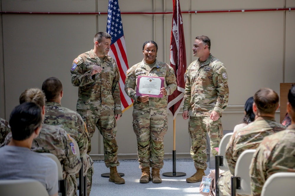 A military awards ceremony is taking place in what appears to be a hangar or indoor military facility. A female soldier in the center is holding a certificate/award while flanked by two male soldiers, with an audience of uniformed personnel seated on both sides. The American flag and a U.S. Army unit guidon are displayed in the background.