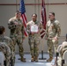 A military awards ceremony is taking place in what appears to be a hangar or indoor military facility. A female soldier in the center is holding a certificate/award while flanked by two male soldiers, with an audience of uniformed personnel seated on both sides. The American flag and a U.S. Army unit guidon are displayed in the background.