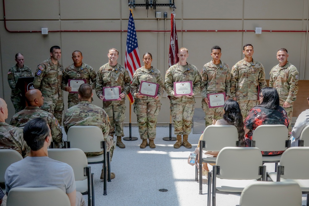 A military awards or recognition ceremony is taking place indoors, likely in a military installation hall or gymnasium. Several soldiers in Army Combat Uniforms (ACU/OCP pattern) are standing in a line holding certificates or award documents with red/maroon folders. An audience of military personnel and civilians is seated facing them.