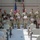 BACH Conducts Commanders Monthly Awards Ceremony A military awards or recognition ceremony is taking place indoors, likely in a military installation hall or gymnasium. Several soldiers in Army Combat Uniforms (ACU/OCP pattern) are standing in a line holding certificates or award documents with red/maroon folders. An audience of military personnel and civilians is seated facing them.