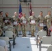 A military awards or recognition ceremony is taking place indoors, likely in a military installation hall or gymnasium. Several soldiers in Army Combat Uniforms (ACU/OCP pattern) are standing in a line holding certificates or award documents with red/maroon folders. An audience of military personnel and civilians is seated facing them.