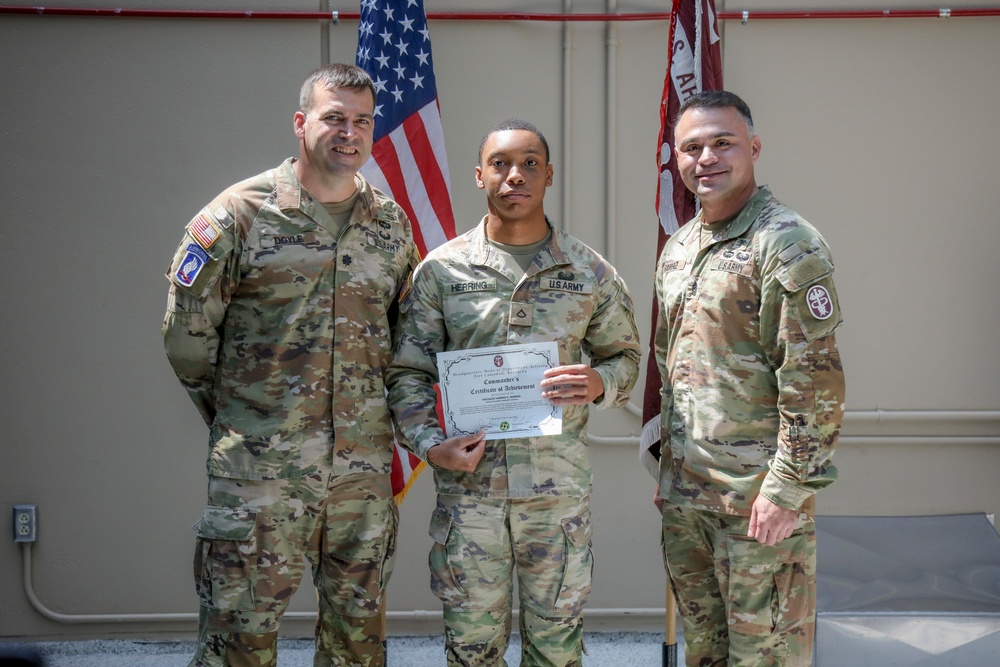 Three U.S. Army soldiers are posed together in what appears to be a military awards ceremony. The center soldier, identified by nametape as 'HERRING,' is holding a Commander's Certificate of Achievement. Two flanking soldiers, identified by nametapes as 'DOYLE' (left) and a partially visible name on the right, appear to be presenting or witnessing the award.