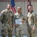 Three U.S. Army soldiers are posed together in what appears to be a military awards ceremony. The center soldier, identified by nametape as 'HERRING,' is holding a Commander's Certificate of Achievement. Two flanking soldiers, identified by nametapes as 'DOYLE' (left) and a partially visible name on the right, appear to be presenting or witnessing the award.