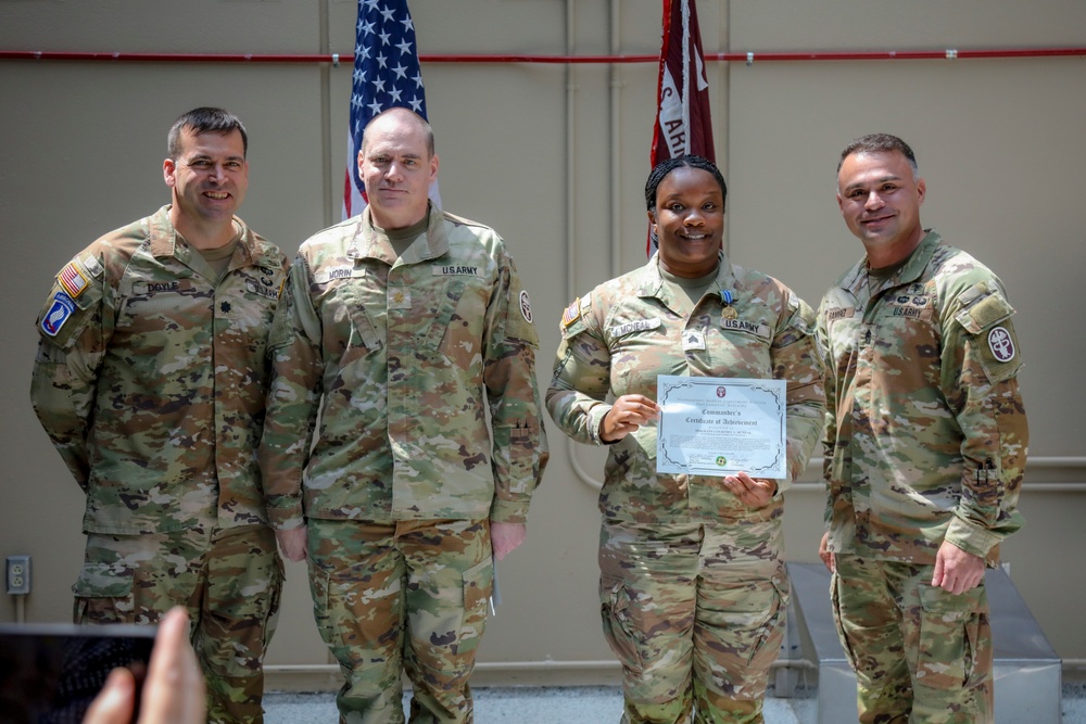 Four U.S. Army soldiers in OCP (Operational Camouflage Pattern) uniforms pose for a photo during a military awards ceremony. One female soldier holds a 'Commander's Certificate of Achievement' and appears to be wearing a medal, indicating she is the recipient of the award. The group is standing in front of American and unit flags in what appears to be a military facility.
