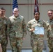 Four U.S. Army soldiers in OCP (Operational Camouflage Pattern) uniforms pose for a photo during a military awards ceremony. One female soldier holds a 'Commander's Certificate of Achievement' and appears to be wearing a medal, indicating she is the recipient of the award. The group is standing in front of American and unit flags in what appears to be a military facility.