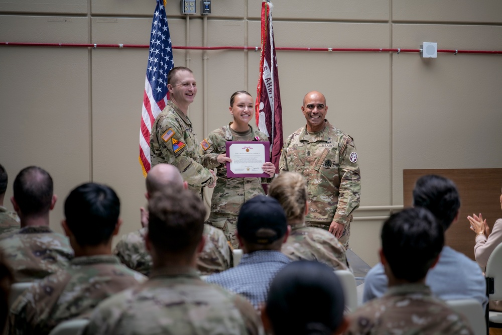 A military award ceremony is taking place in an indoor venue, where a female soldier is receiving a certificate or commendation document from two higher-ranking soldiers. She is flanked by two male soldiers in U.S. Army OCP (Operational Camouflage Pattern) uniforms, all smiling for the occasion. An audience of uniformed and civilian personnel is seated in the foreground observing the ceremony.