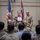 BACH Conducts Commanders Monthly Awards Ceremony A military award ceremony is taking place in an indoor venue, where a female soldier is receiving a certificate or commendation document from two higher-ranking soldiers. She is flanked by two male soldiers in U.S. Army OCP (Operational Camouflage Pattern) uniforms, all smiling for the occasion. An audience of uniformed and civilian personnel is seated in the foreground observing the ceremony.