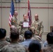 A military award ceremony is taking place in an indoor venue, where a female soldier is receiving a certificate or commendation document from two higher-ranking soldiers. She is flanked by two male soldiers in U.S. Army OCP (Operational Camouflage Pattern) uniforms, all smiling for the occasion. An audience of uniformed and civilian personnel is seated in the foreground observing the ceremony.