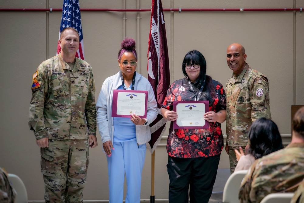 Two civilian employees are being recognized at a U.S. Army awards ceremony, each holding a certificate/award. They are flanked by two U.S. Army soldiers in OCP (Operational Camouflage Pattern) uniforms. The event appears to take place in a military facility with an American flag and a unit guidon visible in the background.