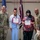 BACH Conducts Commanders Monthly Awards Ceremony Two civilian employees are being recognized at a U.S. Army awards ceremony, each holding a certificate/award. They are flanked by two U.S. Army soldiers in OCP (Operational Camouflage Pattern) uniforms. The event appears to take place in a military facility with an American flag and a unit guidon visible in the background.