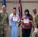 Two civilian employees are being recognized at a U.S. Army awards ceremony, each holding a certificate/award. They are flanked by two U.S. Army soldiers in OCP (Operational Camouflage Pattern) uniforms. The event appears to take place in a military facility with an American flag and a unit guidon visible in the background.