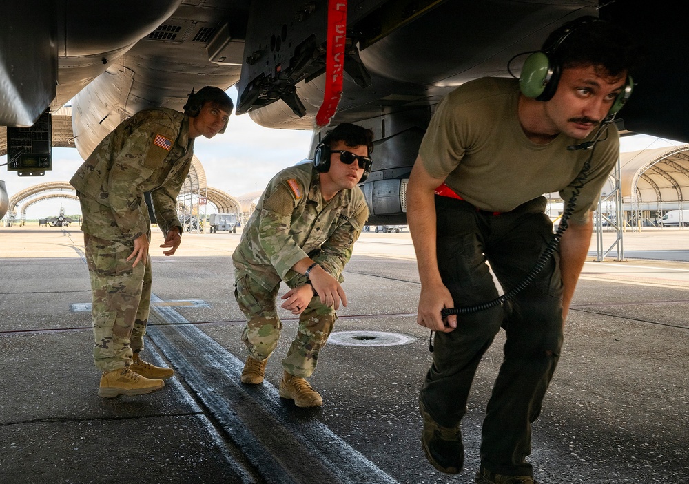 Three military personnel are conducting a pre-flight or maintenance inspection beneath the fuselage of a military aircraft on a flight line tarmac. Two individuals are wearing camouflage uniforms with headsets, while a third wears an olive t-shirt and black pants with a communication headset. The background shows additional aircraft shelters and parked aircraft on what appears to be a military airbase.
