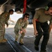 Three military personnel are conducting a pre-flight or maintenance inspection beneath the fuselage of a military aircraft on a flight line tarmac. Two individuals are wearing camouflage uniforms with headsets, while a third wears an olive t-shirt and black pants with a communication headset. The background shows additional aircraft shelters and parked aircraft on what appears to be a military airbase.