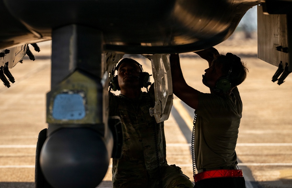 Two military personnel in uniform are performing maintenance or inspection work on the underside of a military aircraft on a flight line or tarmac. Both individuals are wearing communication headsets and are closely examining aircraft components beneath the fuselage. The image is dramatically backlit, creating strong silhouette contrast against the warm ambient light.