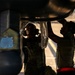 Two military personnel in uniform are performing maintenance or inspection work on the underside of a military aircraft on a flight line or tarmac. Both individuals are wearing communication headsets and are closely examining aircraft components beneath the fuselage. The image is dramatically backlit, creating strong silhouette contrast against the warm ambient light.