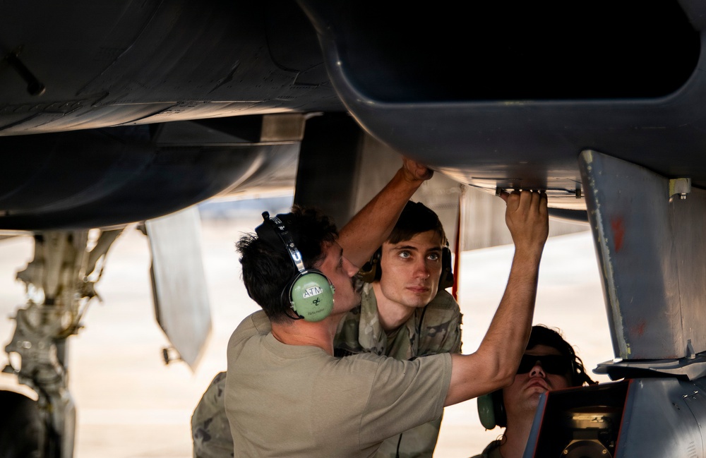 Military aircraft maintenance personnel are working underneath a military jet aircraft, performing what appears to be inspection or repair work on the underside of the fuselage or weapons bay area. At least three individuals are visible, with two actively working on the aircraft components while wearing hearing protection. The setting appears to be an outdoor flight line or tarmac in a warm, arid environment.