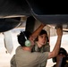 Military aircraft maintenance personnel are working underneath a military jet aircraft, performing what appears to be inspection or repair work on the underside of the fuselage or weapons bay area. At least three individuals are visible, with two actively working on the aircraft components while wearing hearing protection. The setting appears to be an outdoor flight line or tarmac in a warm, arid environment.
