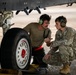 Two military personnel are crouched near the landing gear of an aircraft, appearing to discuss or inspect the wheel assembly. One individual is in civilian-style work clothes with a mustache, while the other wears U.S. Army camouflage (OCP) uniform with an Airborne patch visible on the shoulder. Both are wearing green aviation-style hearing protection headsets.