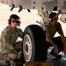 Two military personnel are crouching beneath an aircraft, inspecting the landing gear assembly on a tarmac. The individual on the left is wearing a camouflage uniform with a U.S. flag patch, sunglasses, and green ear protection headset, while the individual on the right wears a plain olive t-shirt with a similar headset. The image appears to depict a pre-flight or maintenance inspection of the aircraft's main landing gear.