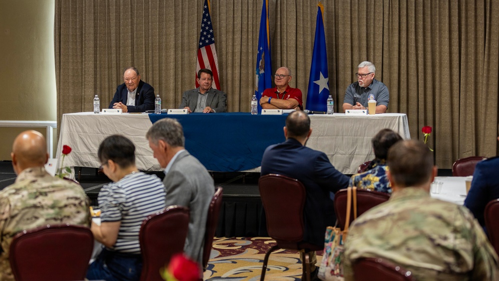A formal panel discussion or briefing is taking place in a conference or banquet room setting. Four panelists are seated at a draped table at the front, with an audience that includes military personnel in camouflage uniforms and civilians seated in rows facing them. Three flags are displayed behind the panel, including the American flag and what appear to be U.S. Air Force-related flags.