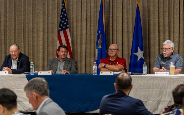 A formal panel discussion or briefing is taking place in a conference or banquet room setting. Four panelists are seated at a draped table at the front, with an audience that includes military personnel in camouflage uniforms and civilians seated in rows facing them. Three flags are displayed behind the panel, including the American flag and what appear to be U.S. Air Force-related flags.