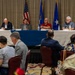 A formal panel discussion or briefing is taking place in a conference or banquet room setting. Four panelists are seated at a draped table at the front, with an audience that includes military personnel in camouflage uniforms and civilians seated in rows facing them. Three flags are displayed behind the panel, including the American flag and what appear to be U.S. Air Force-related flags.