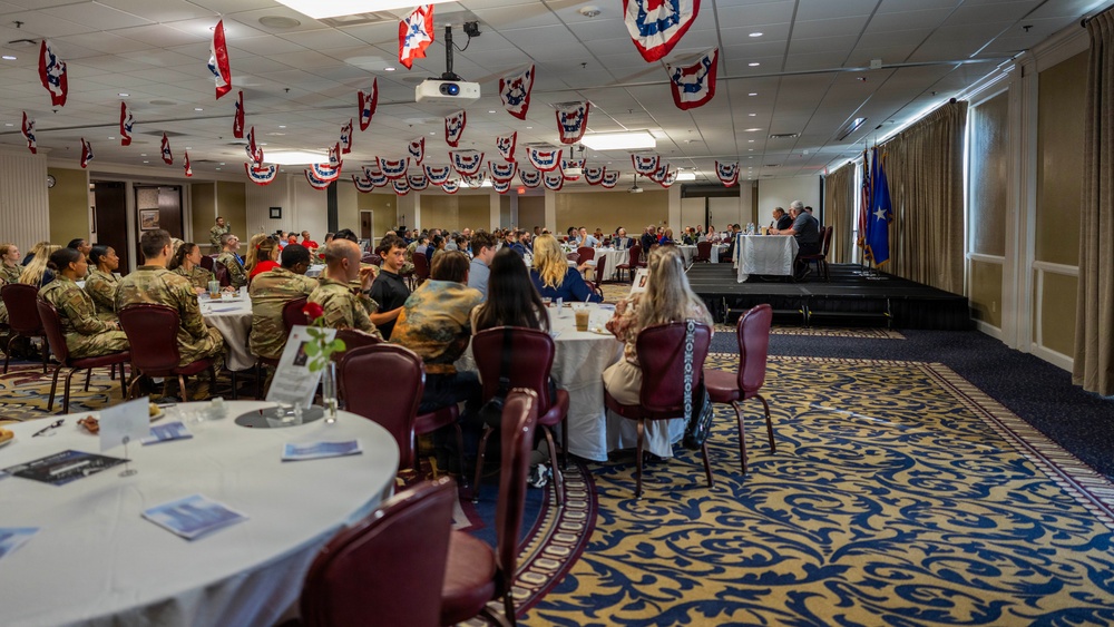A formal military banquet or luncheon is taking place in a large ballroom decorated with red, white, and blue patriotic bunting hanging from the ceiling. Military personnel in Army Combat Uniforms (ACUs) are seated alongside civilians at round tables with white tablecloths. A speaker or presenter appears to be addressing the audience from a raised platform or stage at the far end of the room.