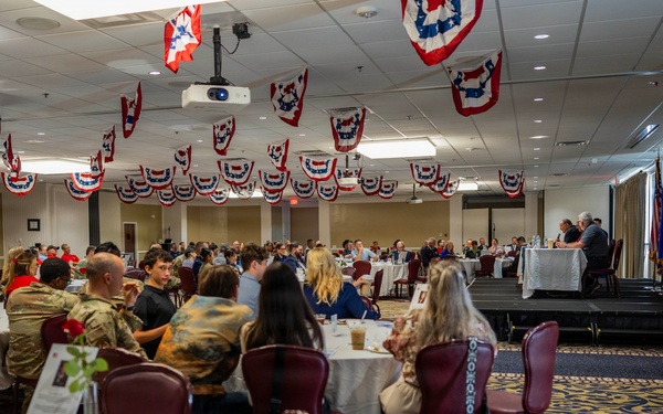 A formal military banquet or luncheon is taking place in a large ballroom decorated with red, white, and blue patriotic bunting hanging from the ceiling. Military personnel in Army Combat Uniforms (ACUs) are seated alongside civilians at round tables with white tablecloths. A speaker or presenter appears to be addressing the audience from a raised platform or stage at the far end of the room.