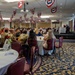 A formal military banquet or luncheon is taking place in a large ballroom decorated with red, white, and blue patriotic bunting hanging from the ceiling. Military personnel in Army Combat Uniforms (ACUs) are seated alongside civilians at round tables with white tablecloths. A speaker or presenter appears to be addressing the audience from a raised platform or stage at the far end of the room.