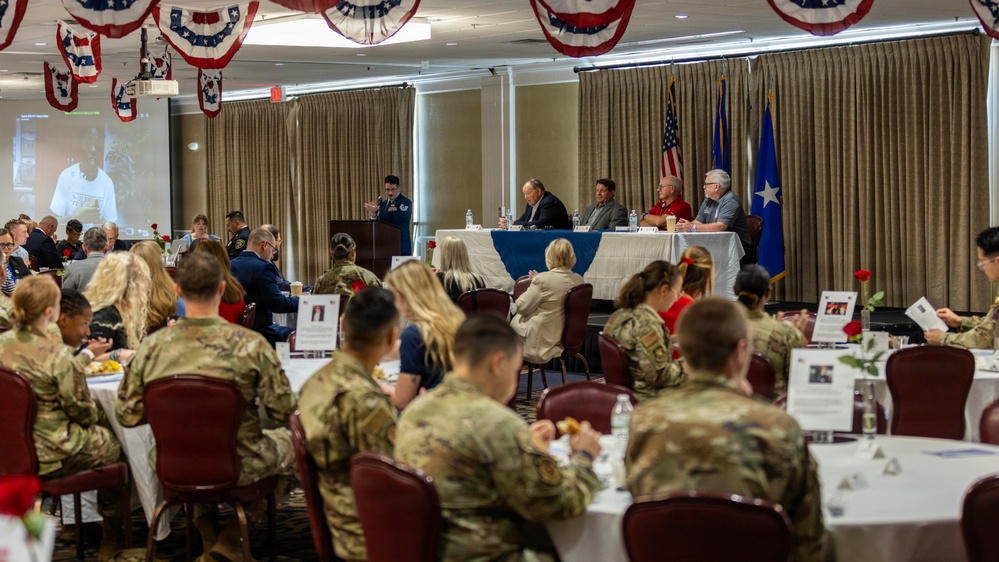 A formal military luncheon or banquet is taking place in a decorated event hall with patriotic red, white, and blue bunting hanging from the ceiling. The majority of attendees are wearing military camouflage uniforms (OCP pattern), seated at round tables with white tablecloths, while a speaker addresses the group from a podium at the front. A panel of individuals is seated at a head table near displayed American and Air Force flags.