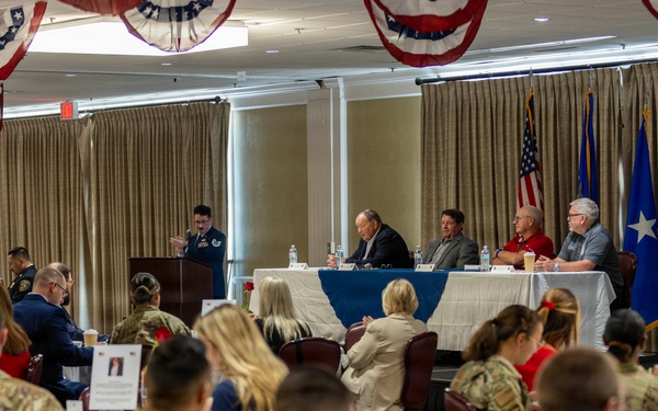 A formal military luncheon or banquet is taking place in a decorated event hall with patriotic red, white, and blue bunting hanging from the ceiling. The majority of attendees are wearing military camouflage uniforms (OCP pattern), seated at round tables with white tablecloths, while a speaker addresses the group from a podium at the front. A panel of individuals is seated at a head table near displayed American and Air Force flags.