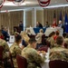 A formal military luncheon or banquet is taking place in a decorated event hall with patriotic red, white, and blue bunting hanging from the ceiling. The majority of attendees are wearing military camouflage uniforms (OCP pattern), seated at round tables with white tablecloths, while a speaker addresses the group from a podium at the front. A panel of individuals is seated at a head table near displayed American and Air Force flags.