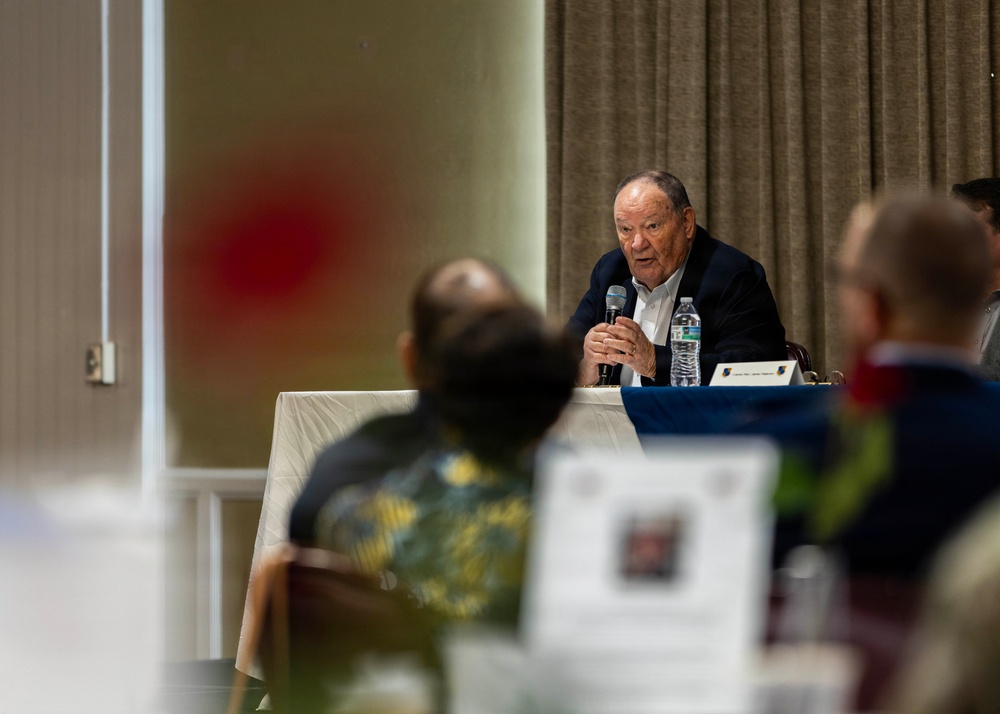 An elderly man in a dark blazer is speaking into a handheld microphone at a panel or conference table, addressing an audience seated in front of him. A water bottle and a name placard are visible on the table before him. The image is shot from behind audience members, with foreground elements intentionally blurred using shallow depth of field.