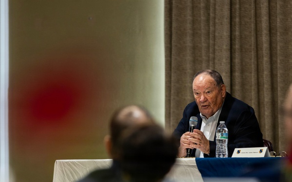 An elderly man in a dark blazer is speaking into a handheld microphone at a panel or conference table, addressing an audience seated in front of him. A water bottle and a name placard are visible on the table before him. The image is shot from behind audience members, with foreground elements intentionally blurred using shallow depth of field.
