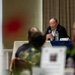 An elderly man in a dark blazer is speaking into a handheld microphone at a panel or conference table, addressing an audience seated in front of him. A water bottle and a name placard are visible on the table before him. The image is shot from behind audience members, with foreground elements intentionally blurred using shallow depth of field.