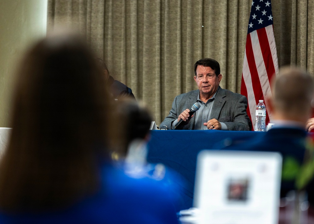A man in a gray blazer is seated at a table with a blue tablecloth, speaking into a handheld microphone at what appears to be a formal meeting or panel discussion. An American flag is visible in the background against draped curtains. The foreground shows audience members, including one using a laptop.