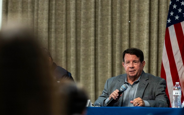 A man in a gray blazer is seated at a table with a blue tablecloth, speaking into a handheld microphone at what appears to be a formal meeting or panel discussion. An American flag is visible in the background against draped curtains. The foreground shows audience members, including one using a laptop.