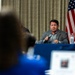 A man in a gray blazer is seated at a table with a blue tablecloth, speaking into a handheld microphone at what appears to be a formal meeting or panel discussion. An American flag is visible in the background against draped curtains. The foreground shows audience members, including one using a laptop.