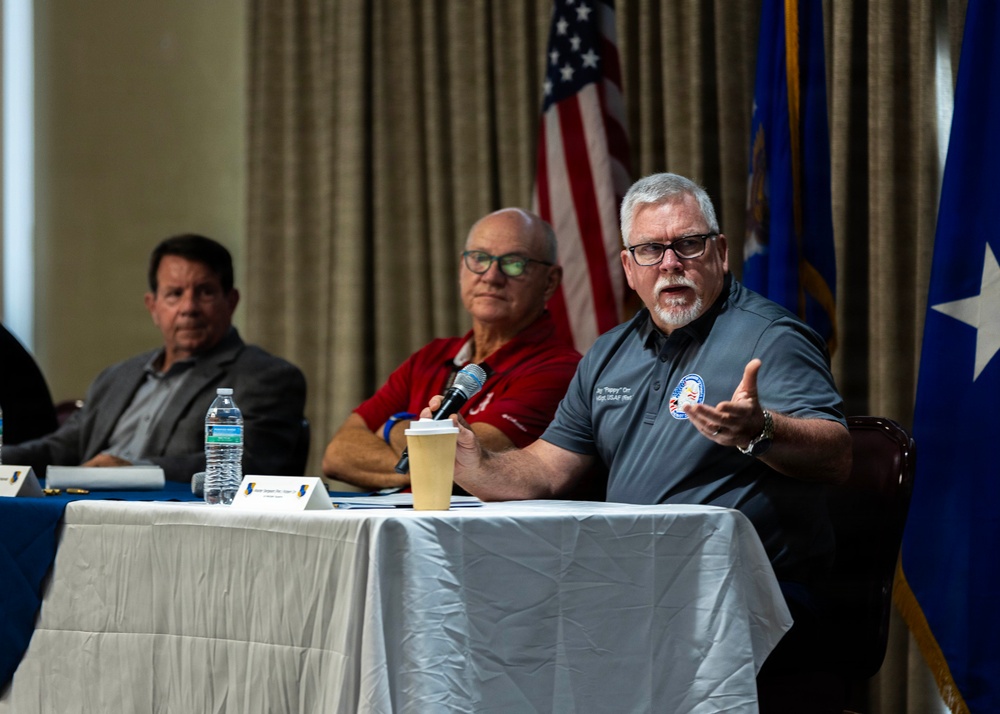 Three men are seated at a panel table during what appears to be an official or military-affiliated forum. The man on the right is actively speaking into a microphone while gesturing with his hand, wearing a gray polo shirt with an embroidered patch. American and Air Force flags are visible in the background, suggesting a U.S. Air Force-related event.