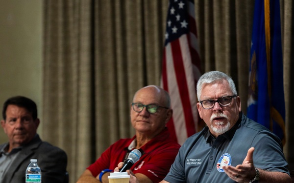 Three men are seated at a panel table during what appears to be an official or military-affiliated forum. The man on the right is actively speaking into a microphone while gesturing with his hand, wearing a gray polo shirt with an embroidered patch. American and Air Force flags are visible in the background, suggesting a U.S. Air Force-related event.