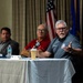 Three men are seated at a panel table during what appears to be an official or military-affiliated forum. The man on the right is actively speaking into a microphone while gesturing with his hand, wearing a gray polo shirt with an embroidered patch. American and Air Force flags are visible in the background, suggesting a U.S. Air Force-related event.