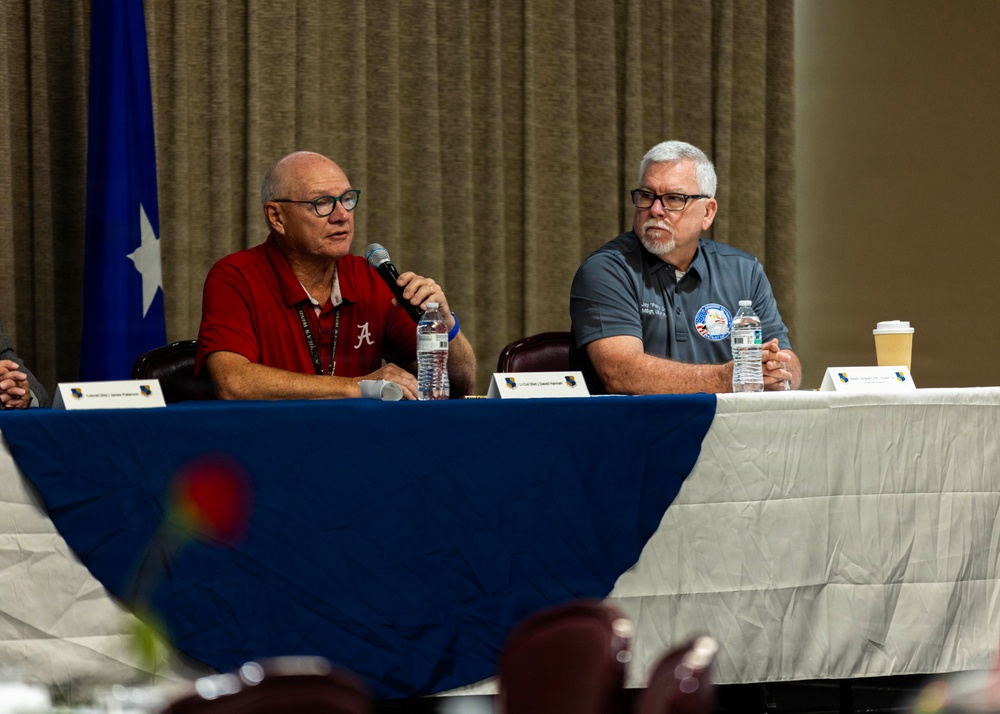 Two older men are seated at a panel table covered with a blue tablecloth, participating in what appears to be a formal discussion or briefing event. The man on the left, wearing a red University of Alabama polo shirt, is speaking into a microphone, while the man on the right in a gray polo shirt listens attentively. Name placards, water bottles, and a military flag are visible, suggesting an official or military-affiliated event.