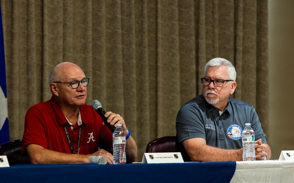 Two older men are seated at a panel table covered with a blue tablecloth, participating in what appears to be a formal discussion or briefing event. The man on the left, wearing a red University of Alabama polo shirt, is speaking into a microphone, while the man on the right in a gray polo shirt listens attentively. Name placards, water bottles, and a military flag are visible, suggesting an official or military-affiliated event.