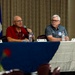 Two older men are seated at a panel table covered with a blue tablecloth, participating in what appears to be a formal discussion or briefing event. The man on the left, wearing a red University of Alabama polo shirt, is speaking into a microphone, while the man on the right in a gray polo shirt listens attentively. Name placards, water bottles, and a military flag are visible, suggesting an official or military-affiliated event.